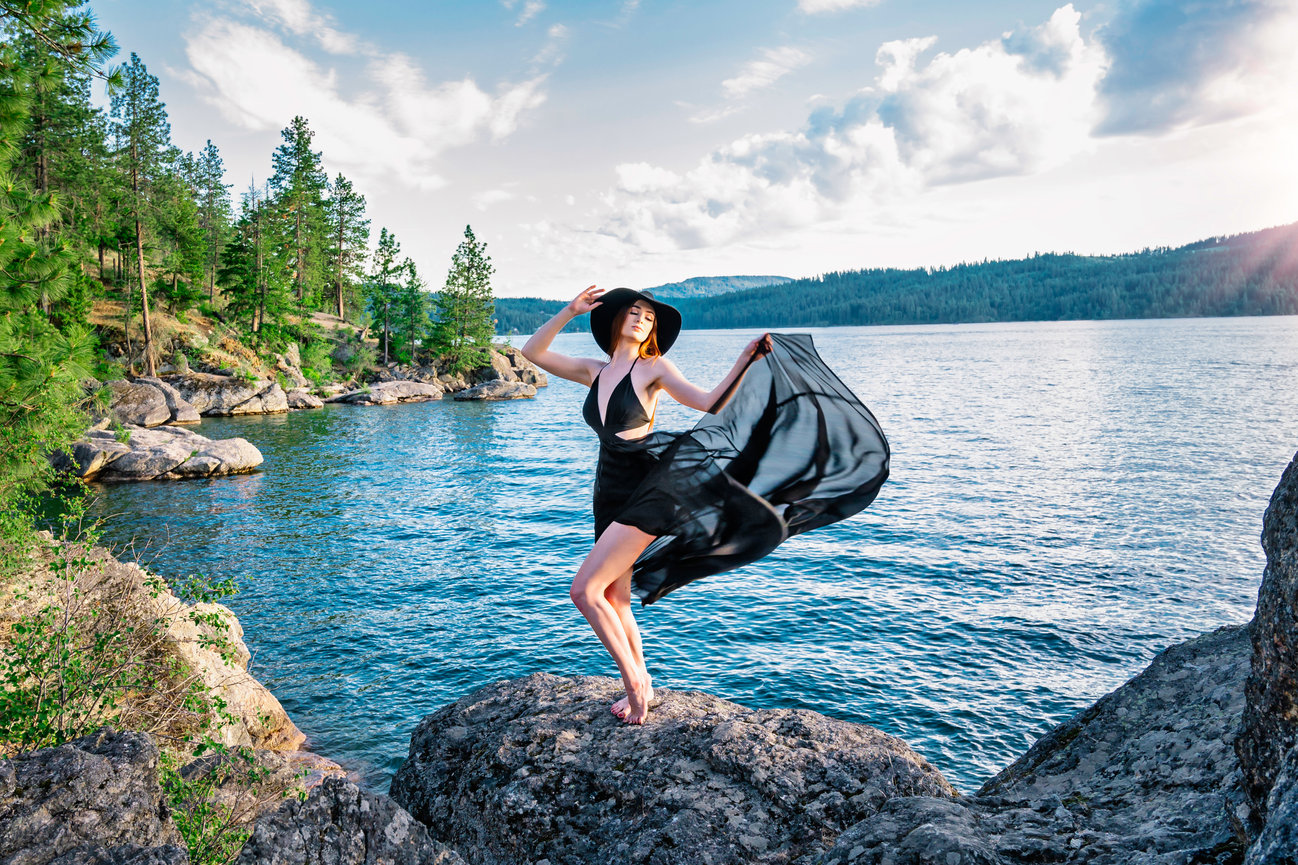 Woman in a black dress and hat poses on a rock by a serene lake, surrounded by trees and mountains.