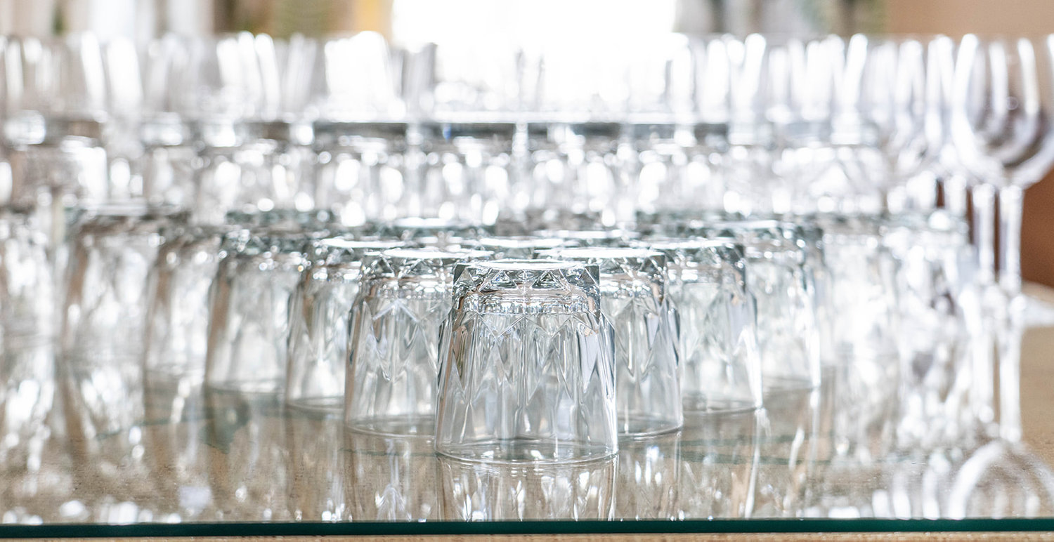 A collection of glassware stacked upside down on a reflective surface, with additional wine glasses in the background.