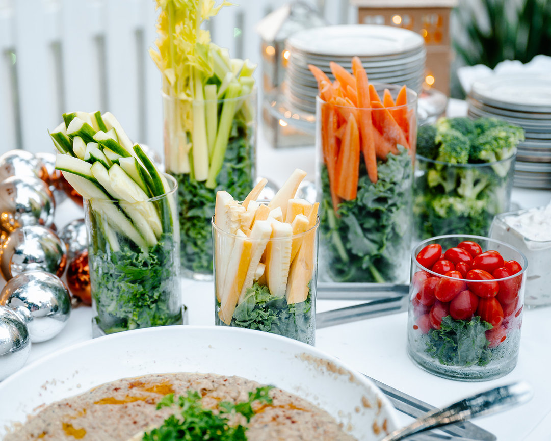 A colorful display of fresh vegetables in glass containers, including celery, cucumber, carrots, broccoli, and cherry tomatoes.