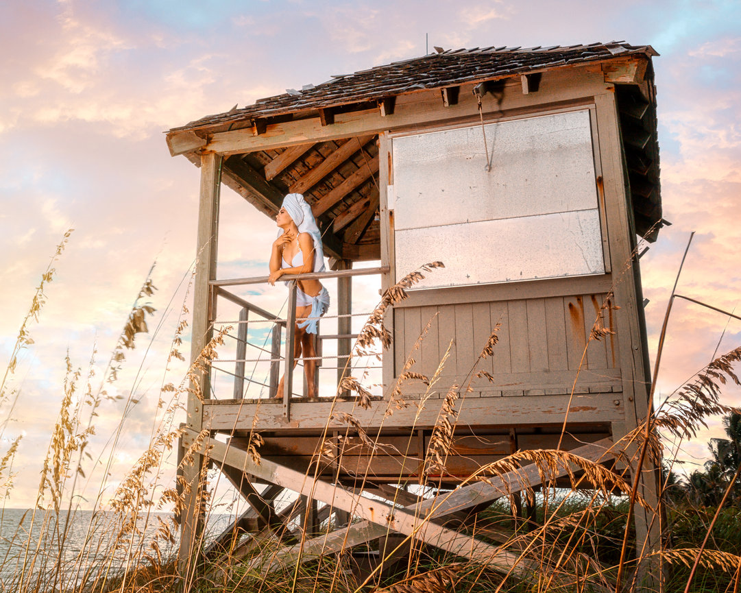 Woman in a towel stands on the balcony of a beach house at sunset, surrounded by tall grass.