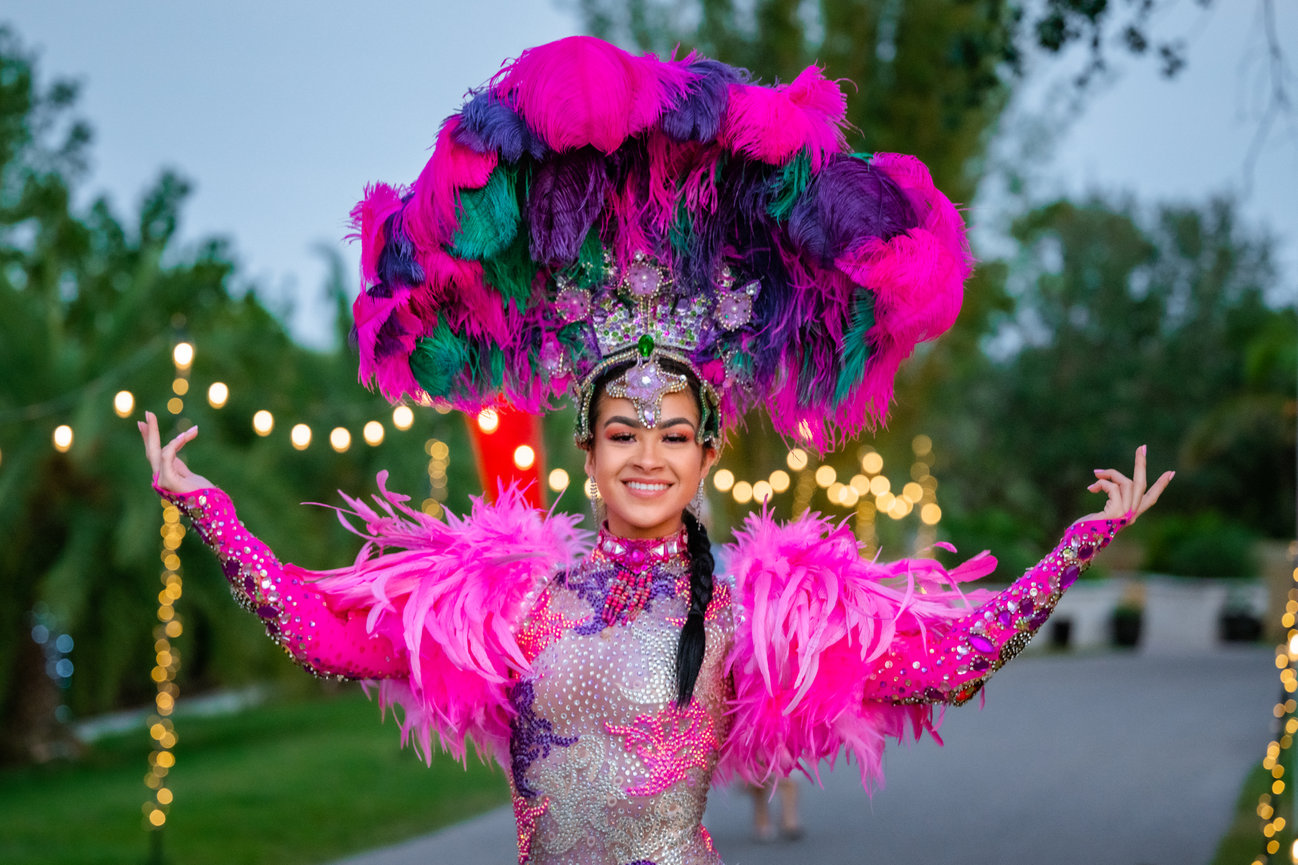 Festival performer wearing a vibrant pink feather costume and colorful headpiece, posing happily outdoors.