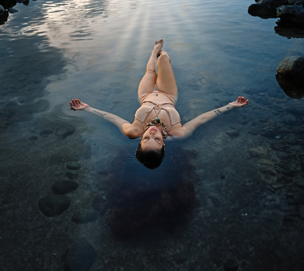 A person in a bikini floats on calm water, with rocks and a reflective sky in the background.