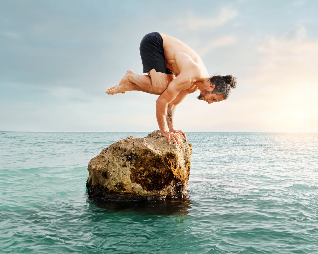 Man performing a balancing pose on a rock in the ocean during sunset.