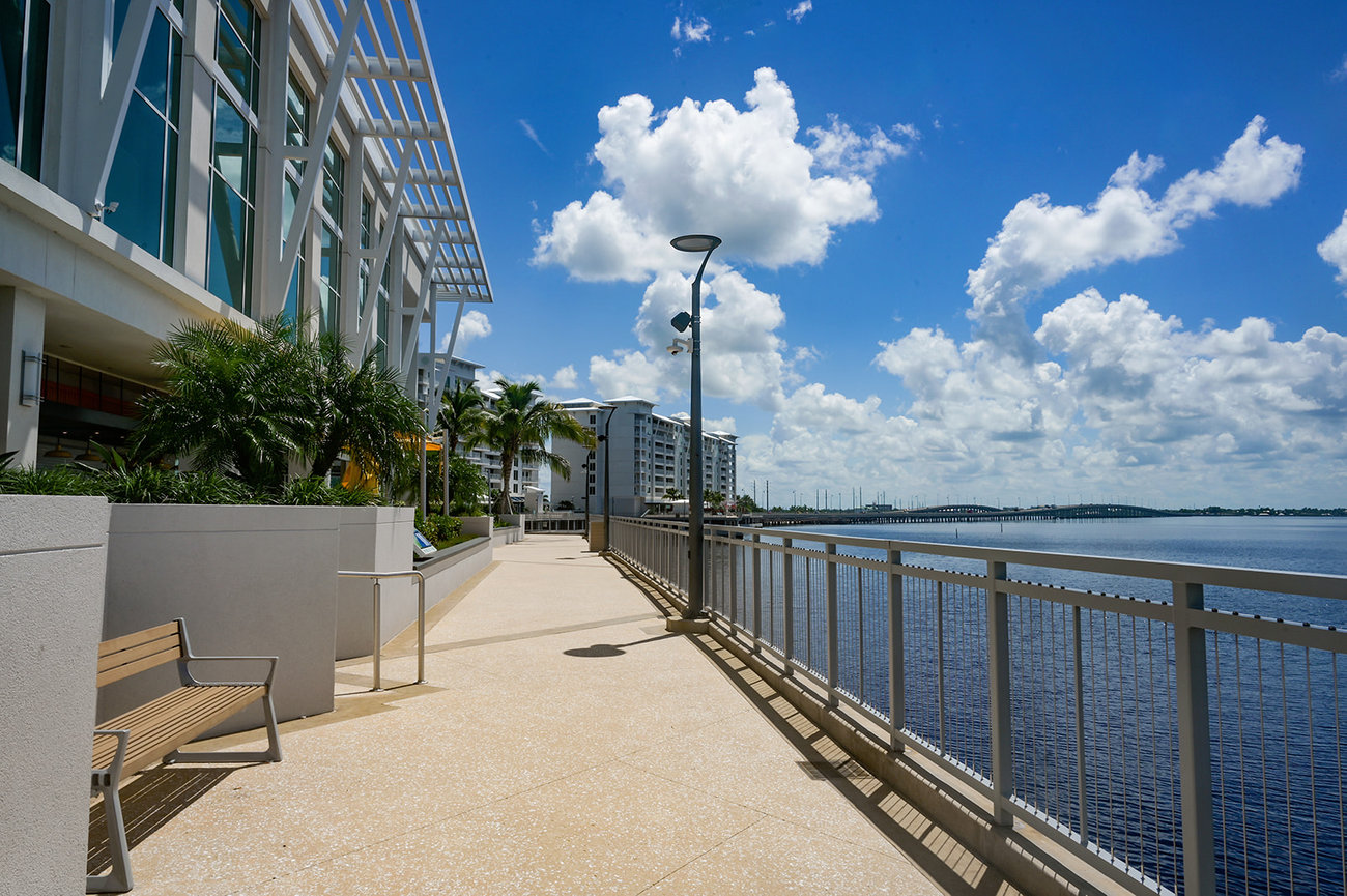 Scenic waterfront walkway with modern buildings, palm trees, and a bright blue sky filled with clouds.