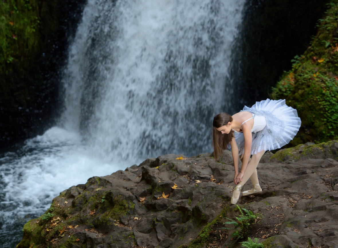 Dancer wearing a tutu lacing her ballet shoe in front of a waterfall at Columbia River Gorge