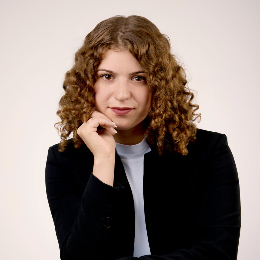 Woman with curly hair in a black blazer, white top, posing thoughtfully against a plain background.