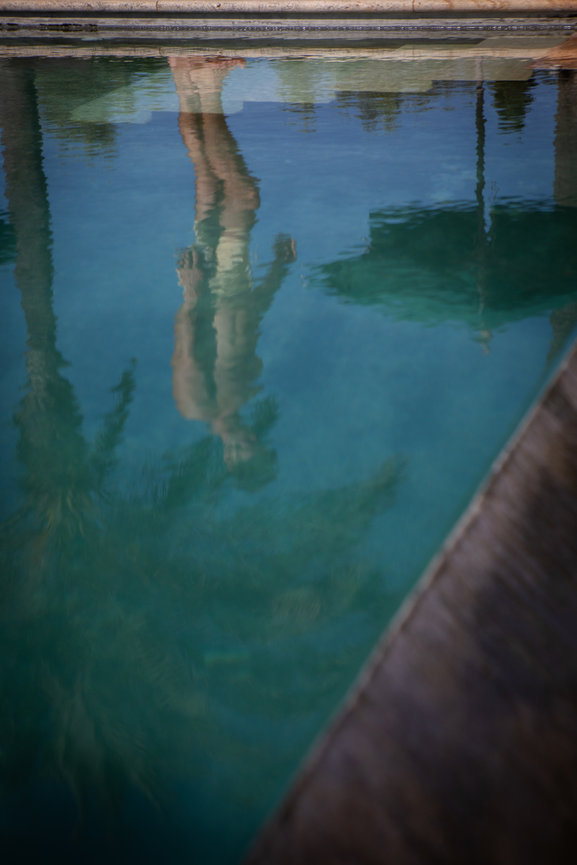 Reflection of a person and palm trees in a swimming pool's calm water.
