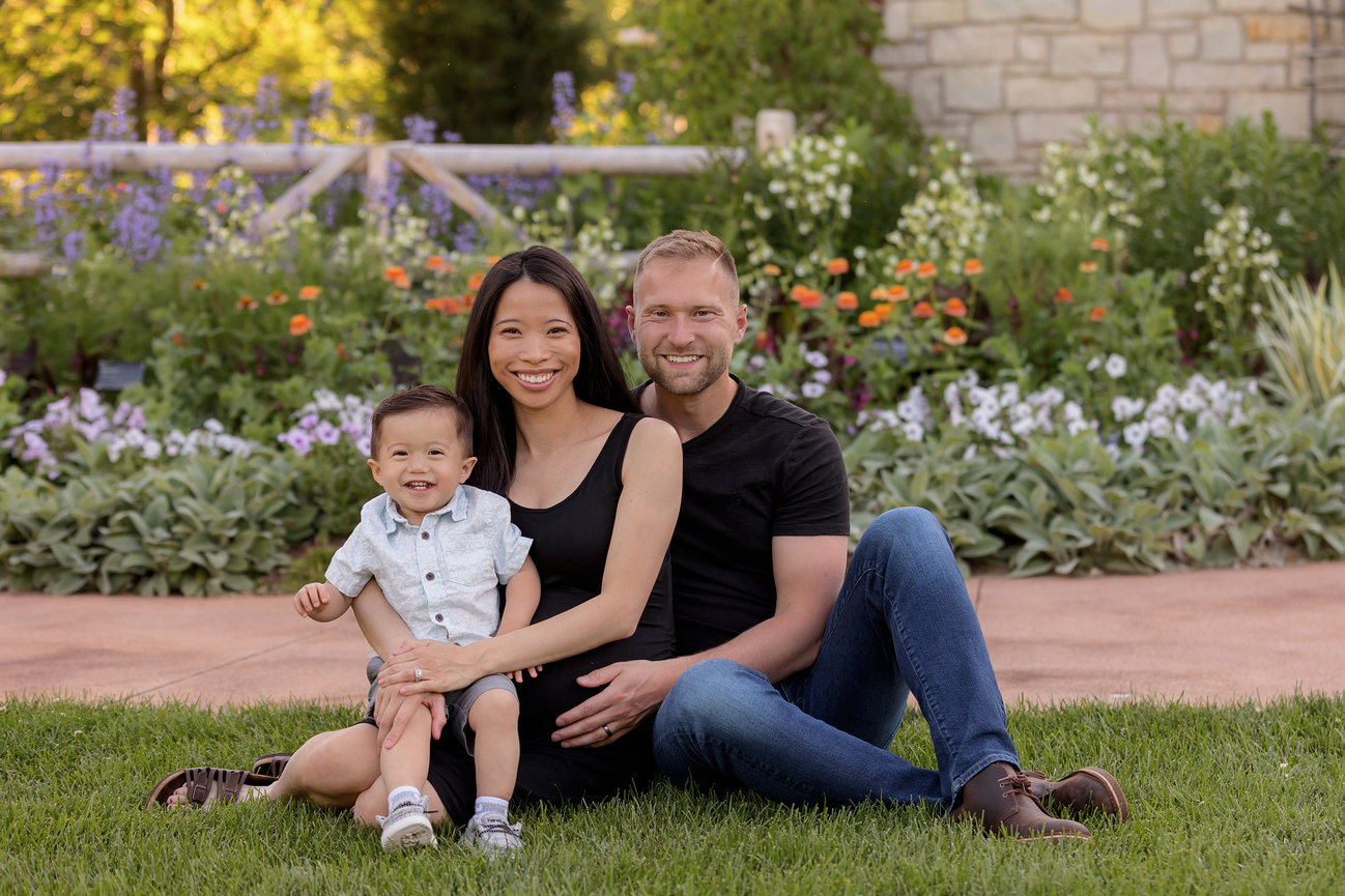 A family of three sitting on grass in a garden with flowers, smiling at the camera.