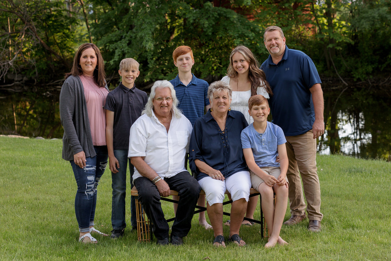 A family poses outside on grass, with trees and water in the background. Two people are seated; others stand around them.
