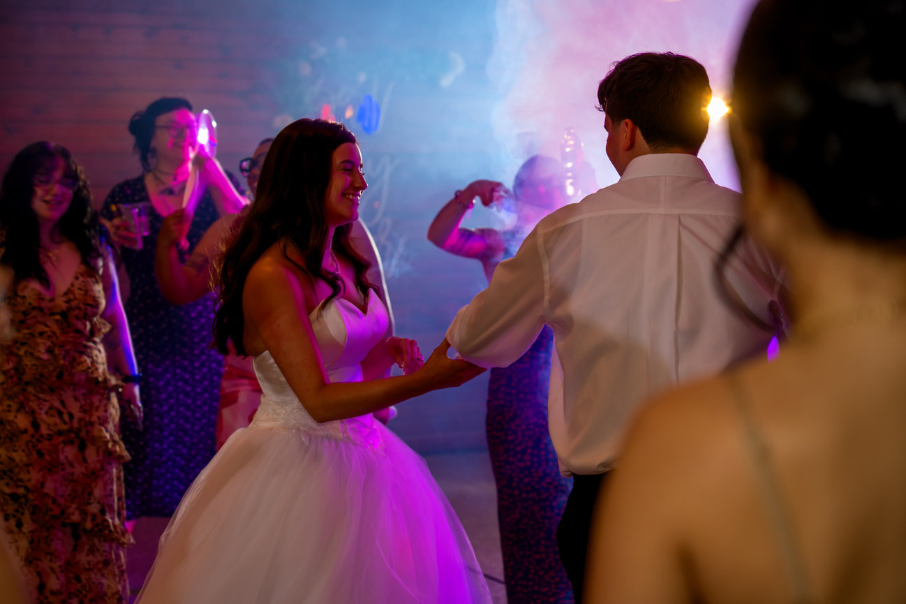 Bride dancing with groom at a lively wedding reception under colorful party lights and smoke.