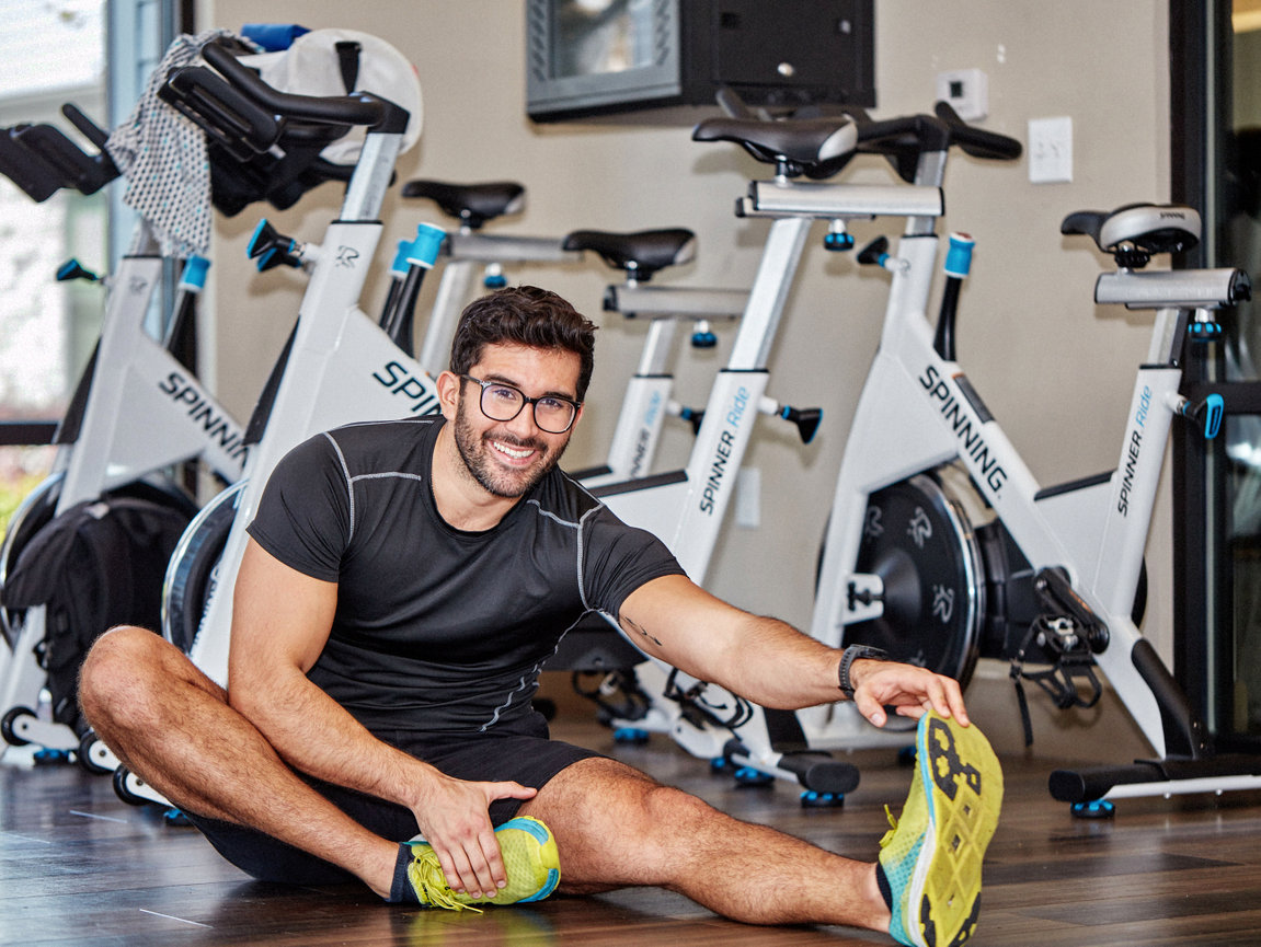 Man stretching on gym floor near spinning bikes, wearing glasses and athletic gear.