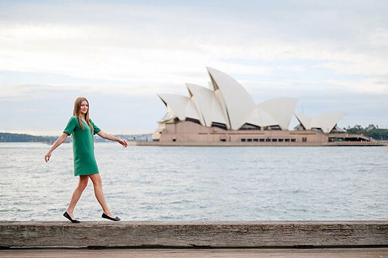 Young woman walking beside Sydney harbour with the Opera House behind her.