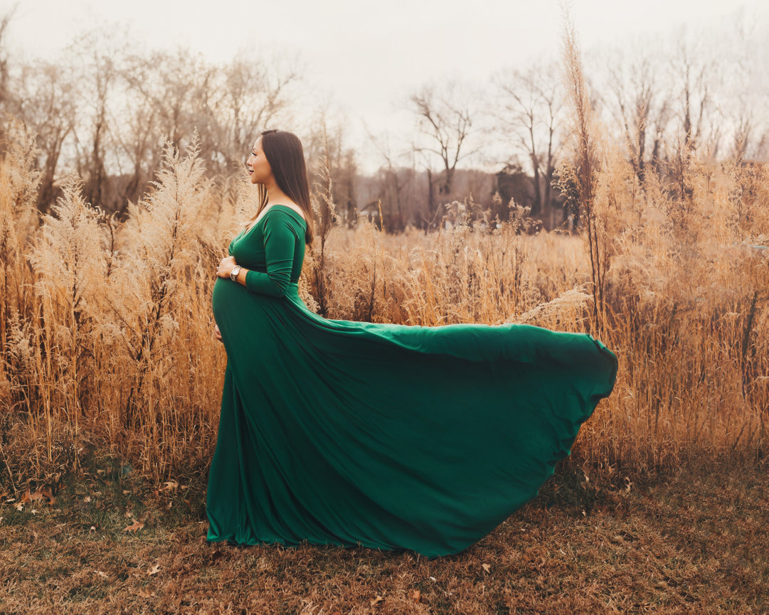 Pregnant woman in a flowing green dress standing in a dry grassy field outdoors