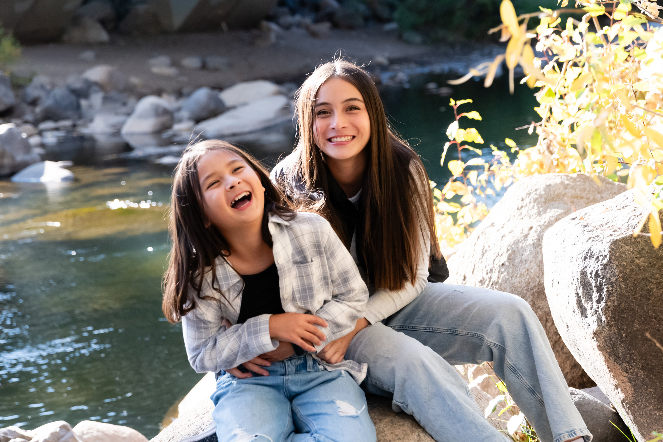 2 girls sitting on a rock by a river hugging and laughing