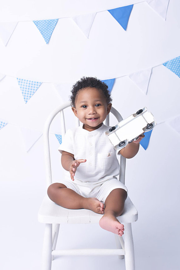 A grinning African-american toddler boy sitting on a white chair holds a white toy truck. There are blue banners behind him.