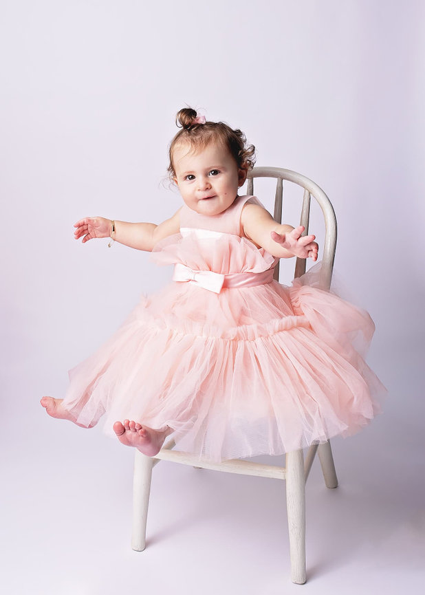 A smiling toddler in pink tulle sits on a white chair with a white background