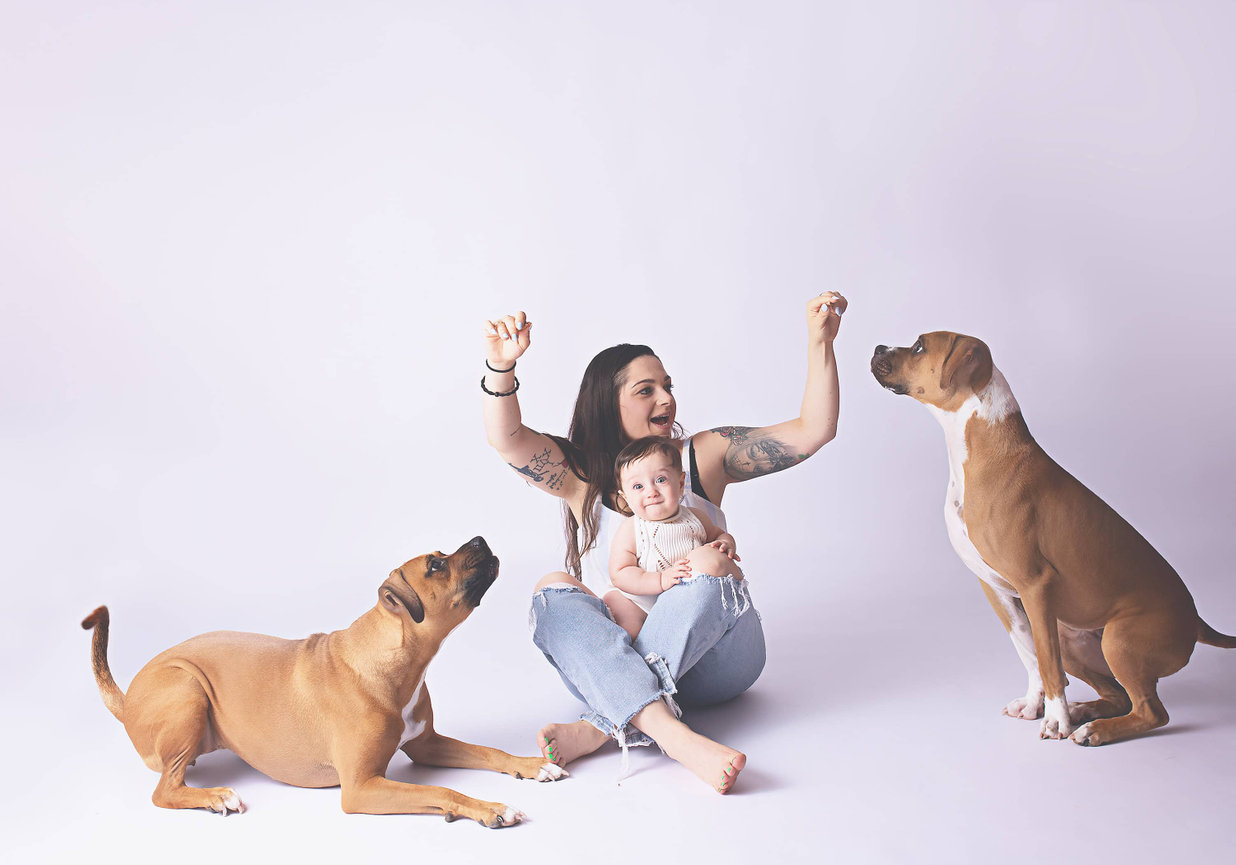 A mom with a baby in her lap holds up treats for two boxer mix dogs. One dog is sitting and one is laying down.