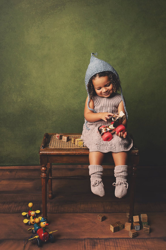 biracial toddler girl in a taupe colored knit dress and blue knit bonnet plays with a vintage toy dog