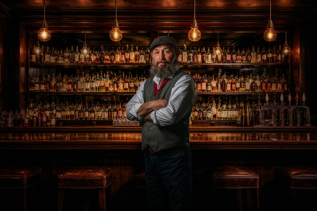 A bearded man in a vest and hat stands confidently at a dimly lit bar with shelves full of bottles.