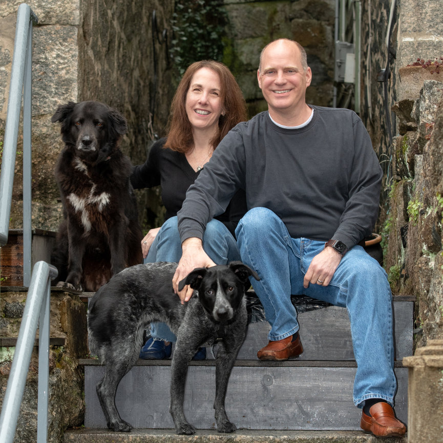 Smiling couple sitting on outdoor steps with two black dogs.