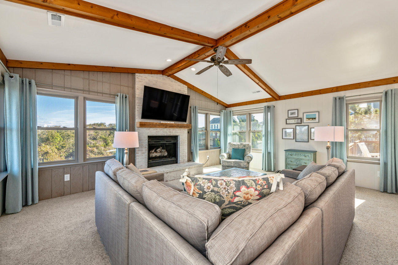 Cozy living room with gray sofas, floral ottoman, fireplace, ceiling fan, TV, and large windows with blue curtains.