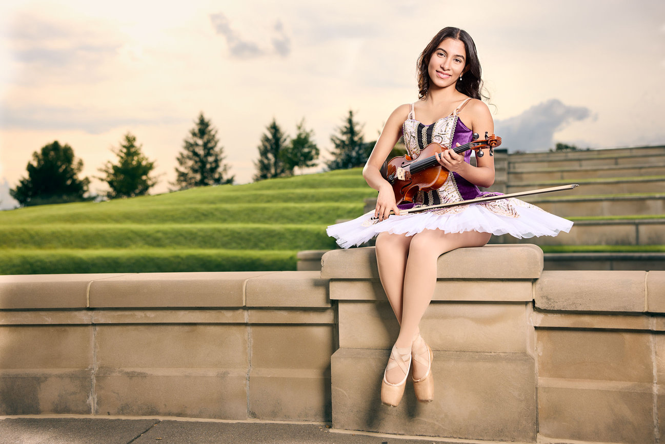 Senior ballerina seated at the Centerpiece in Coxhall Gardens, Carmel, Indiana, wearing a tutu and pointe shoes while holding her violin, photographed by Wahala Portraits.