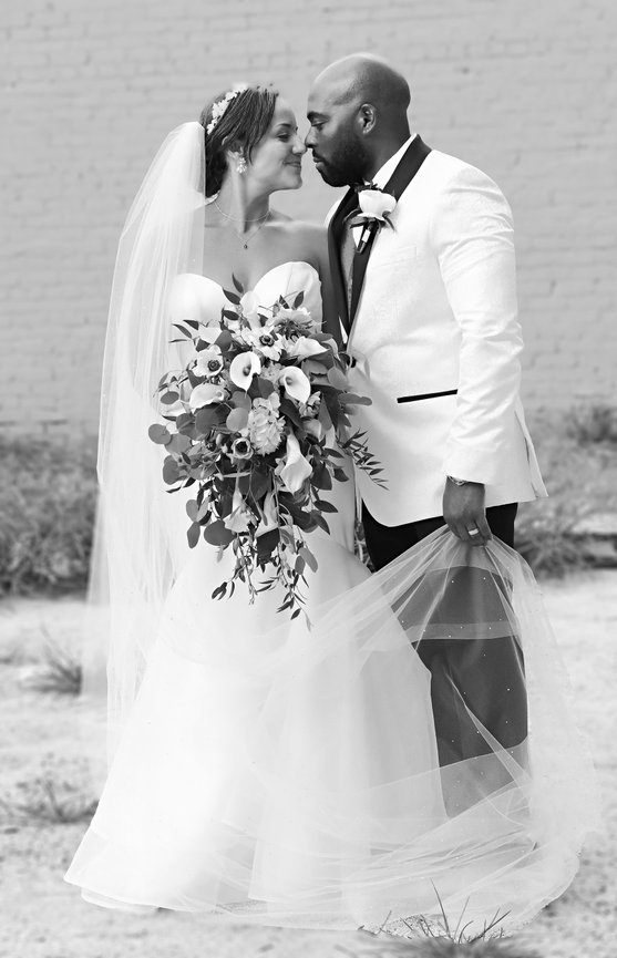 Bride and groom in wedding attire share a kiss, holding a bouquet, in front of a brick wall.