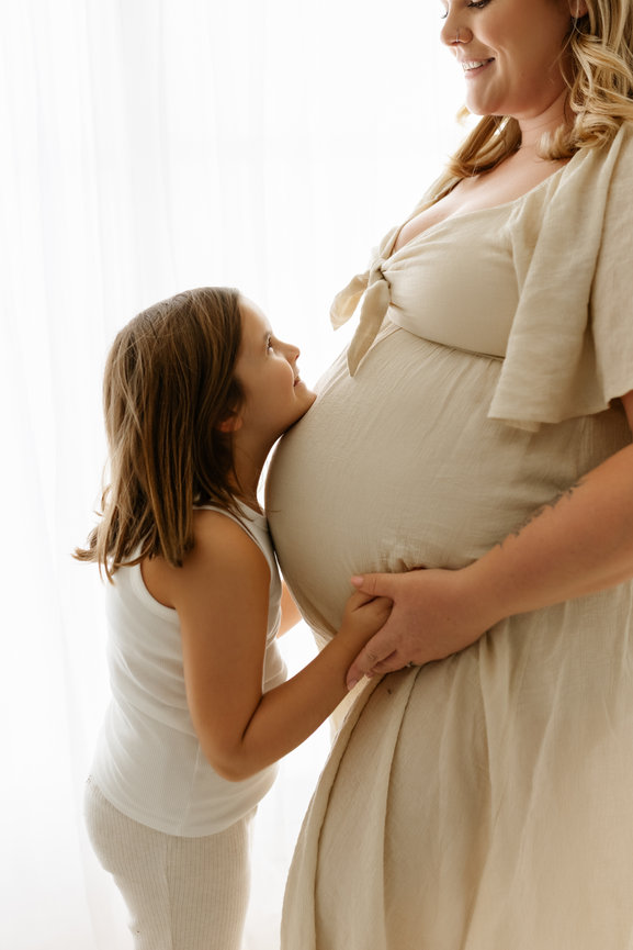 A young girl smiling up at a pregnant woman, touching her belly, in a bright, airy setting at a maternity session in albany ny