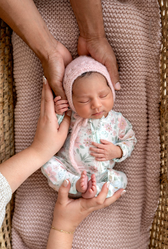 Newborn baby in floral onesie and pink knit bonnet, gently held by hands in a woven basket
