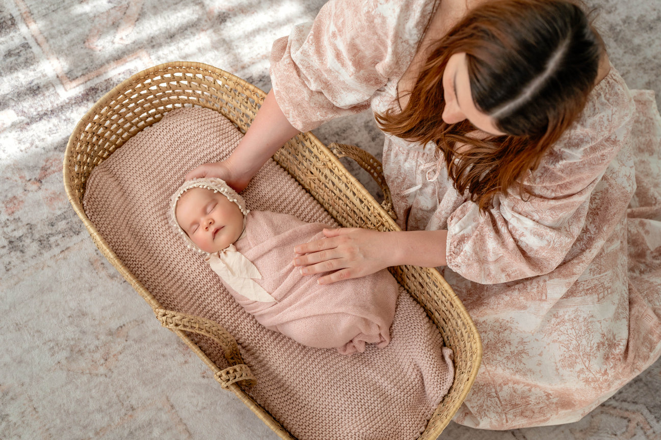 Mother gently holding a sleeping newborn in a woven basket on a soft blanket