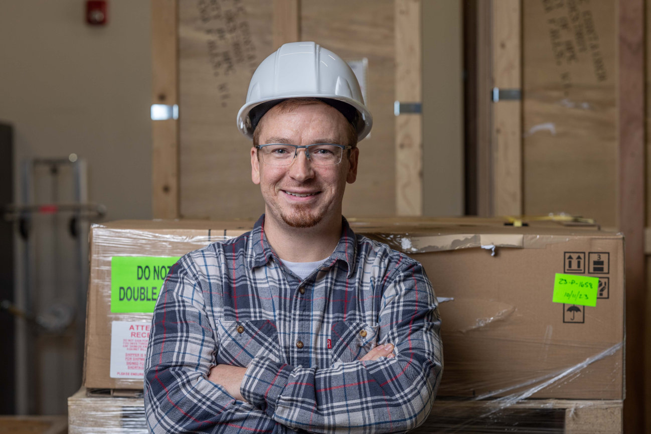 Warehouse worker in a hard hat stands in front of stacked shipping boxes.