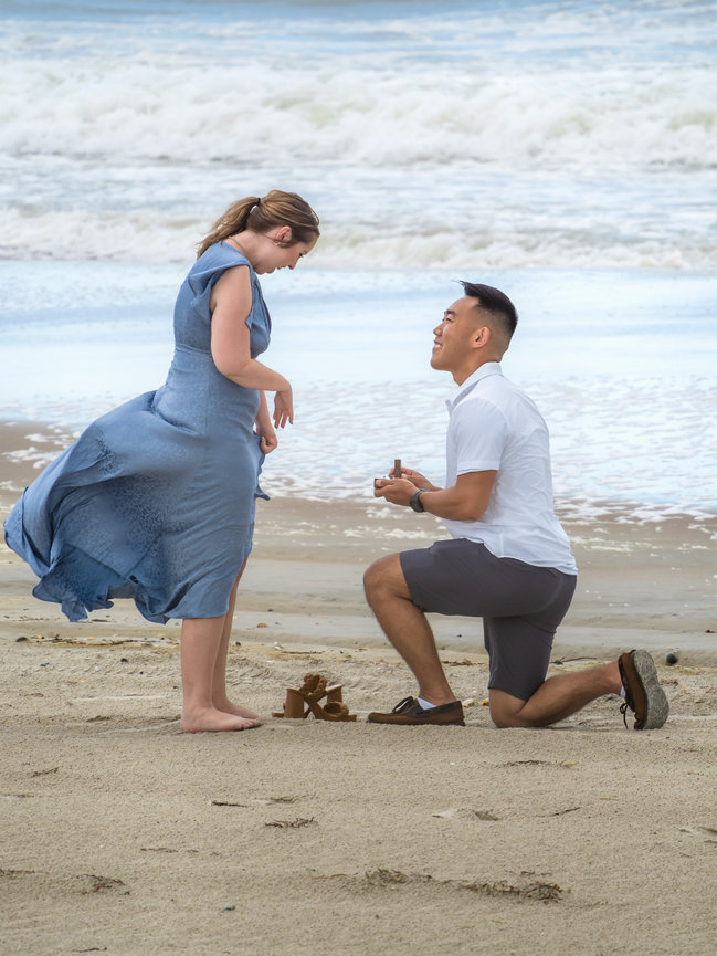 Boyfriend proposes on the beach to girlfriend in a flowing blue dress on one knee with the ring.