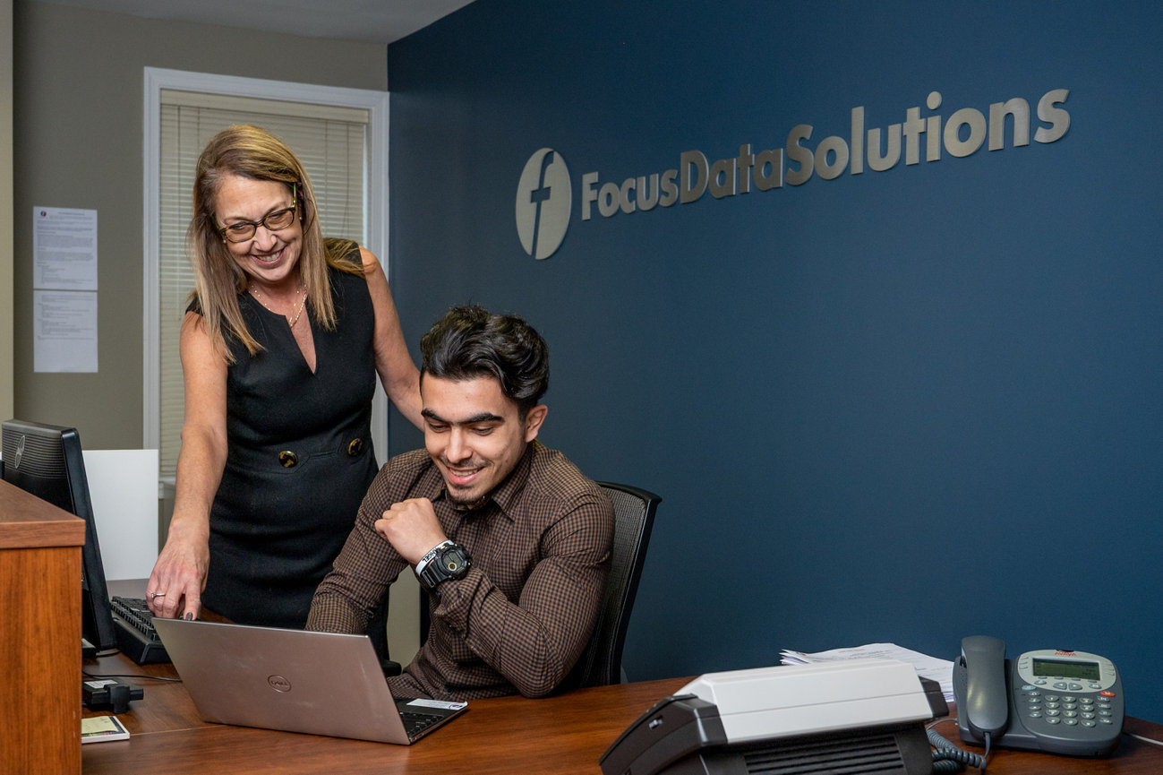 Two coworkers smiling at a laptop in an office with a Focus Data Solutions sign