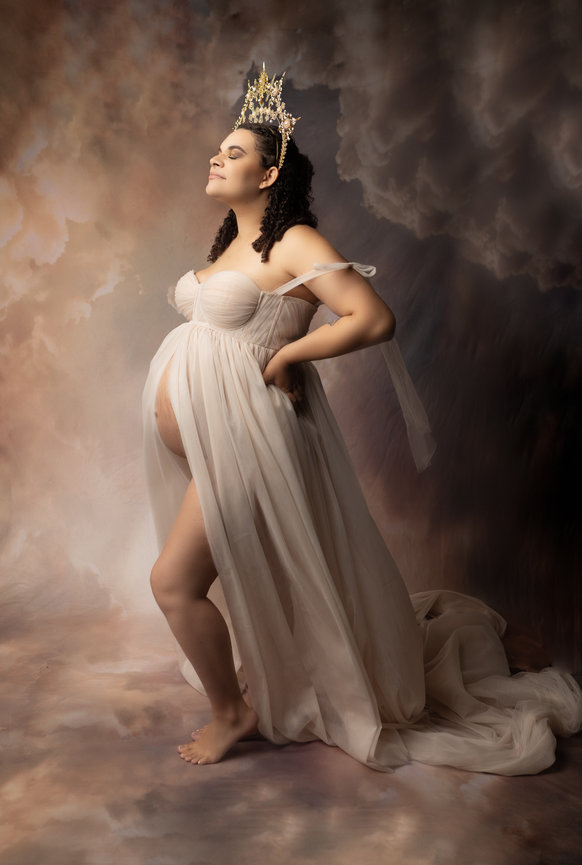 Pregnant woman wearing a flowing gown and crown, stands gracefully in front of a cloud-like background.