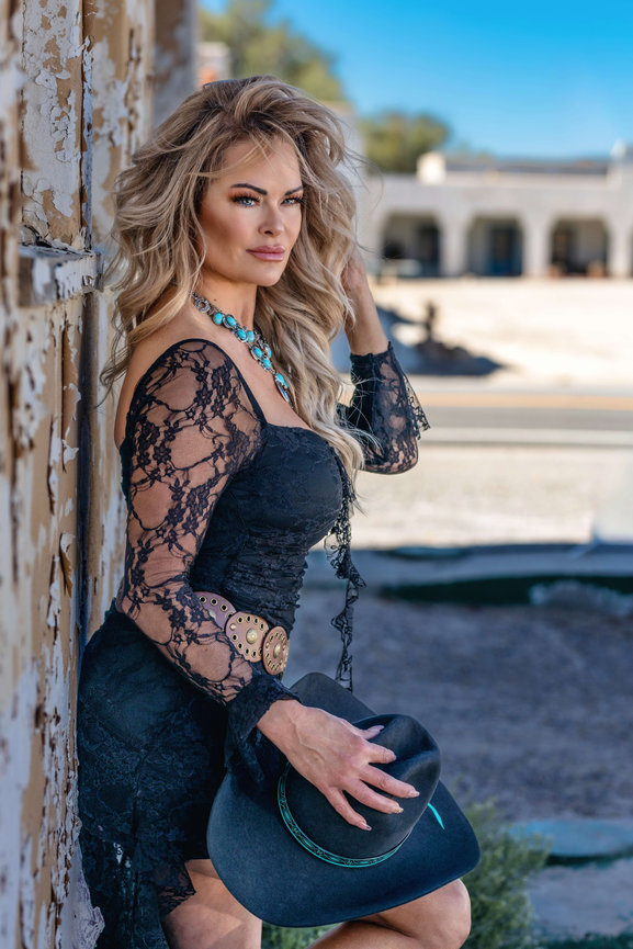 Woman in a black lace dress holding a cowboy hat outdoors by a weathered wall