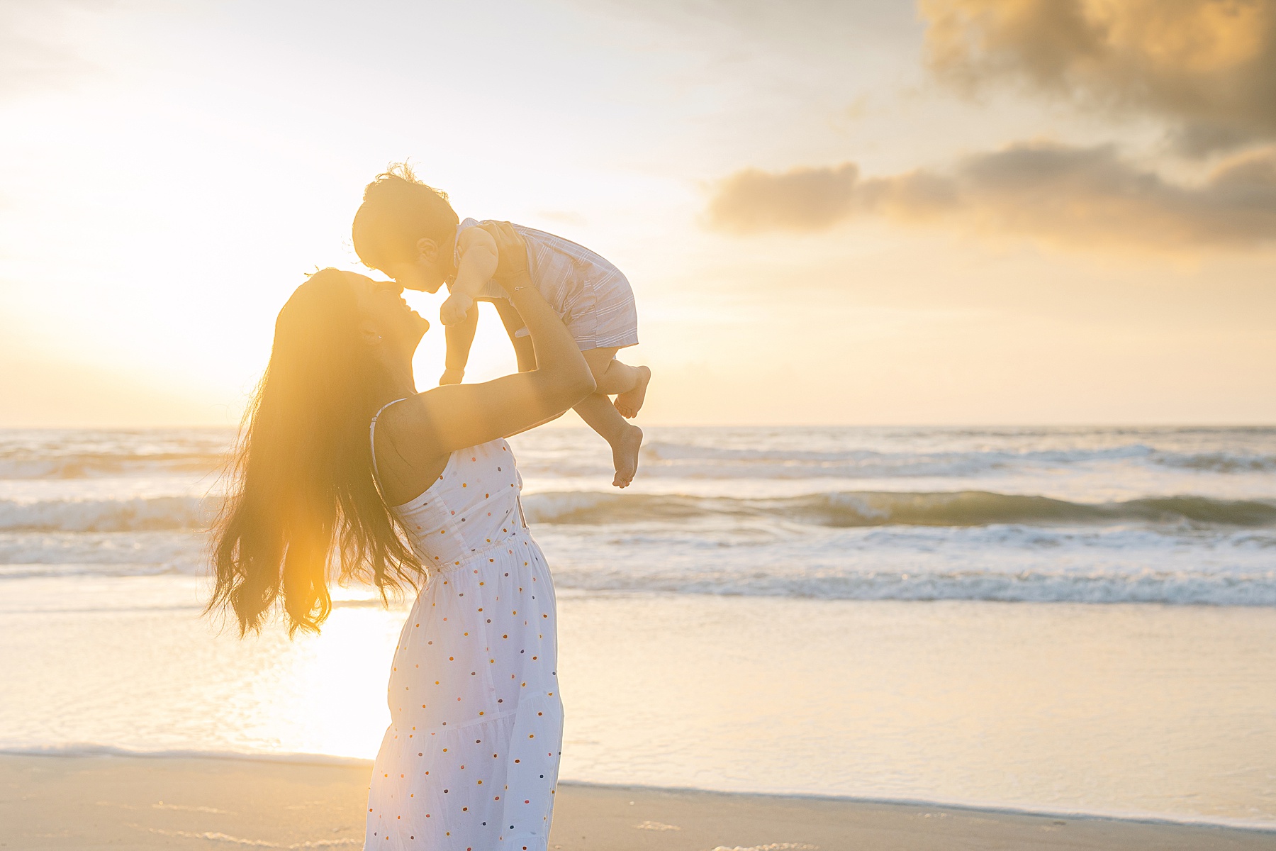 woman in white dress holding baby in backlit sun on the beach at sunrise