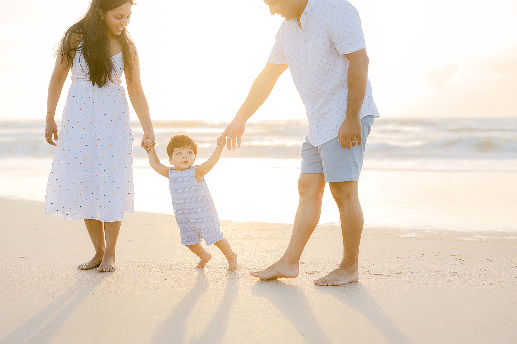 man and woman holding a baby hand while walking on the beach at sunrise