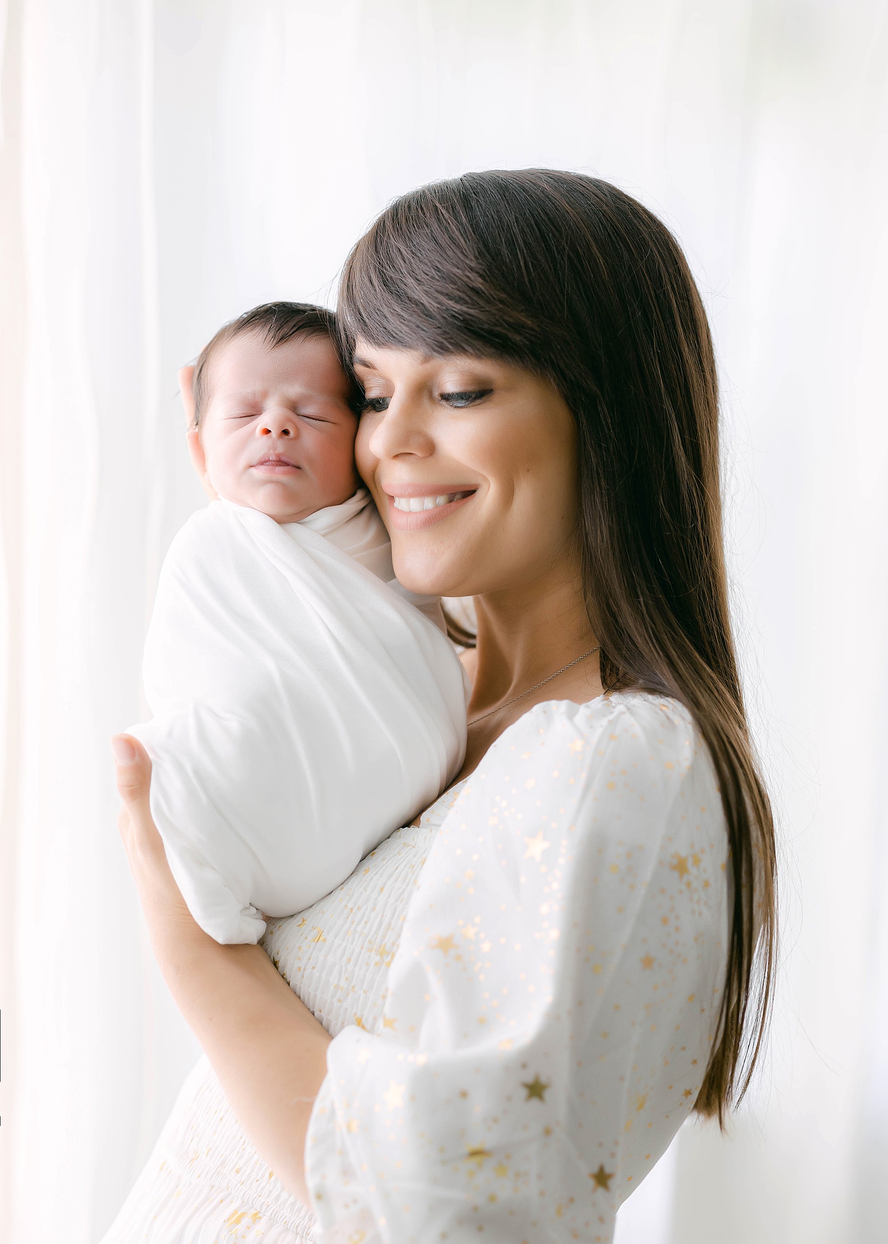woman holding newborn baby in front of window wearing white dress with gold stars