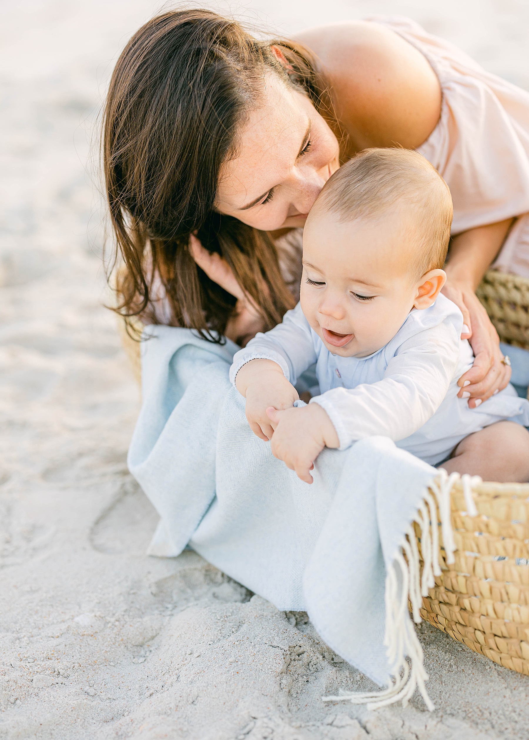 woman smiling kissing baby in basket on the beach sand