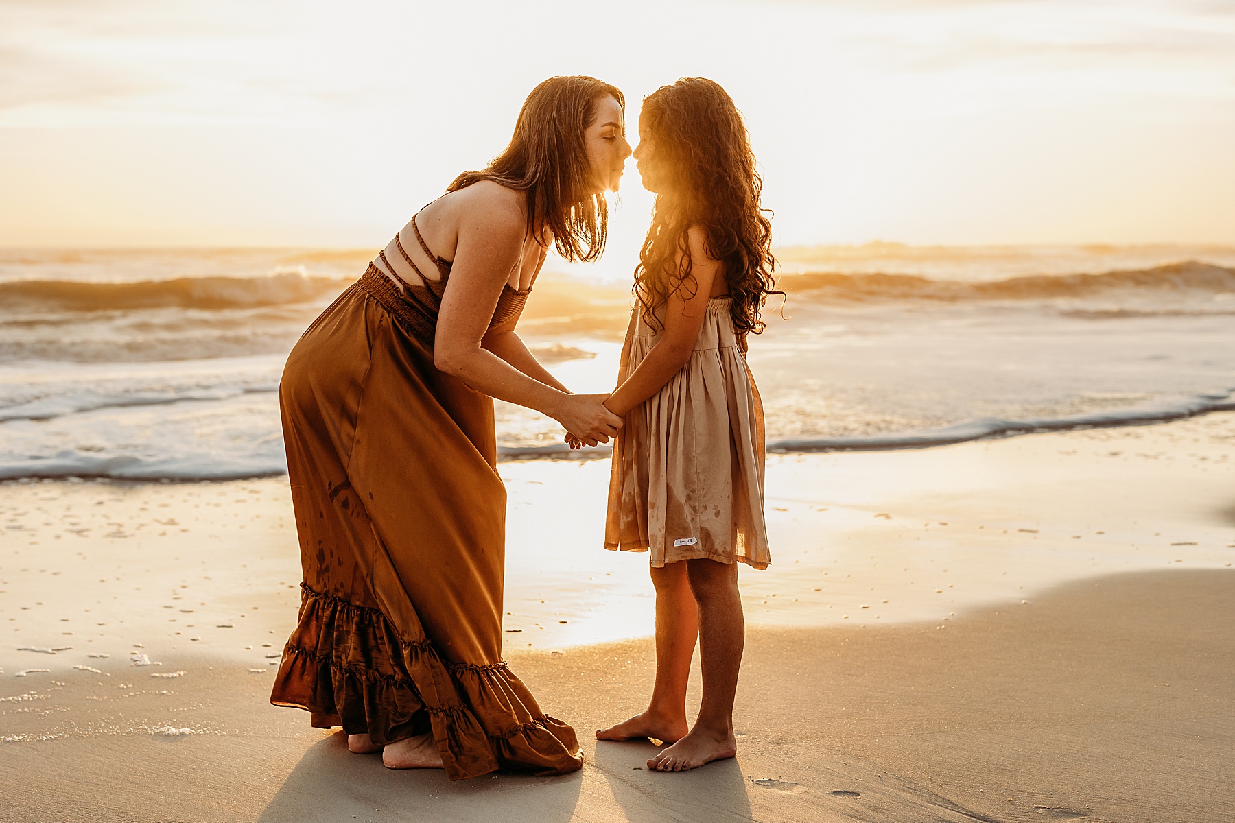 woman kissing little girl on the beach at sunrise wearing warm colors and backlit sunshine