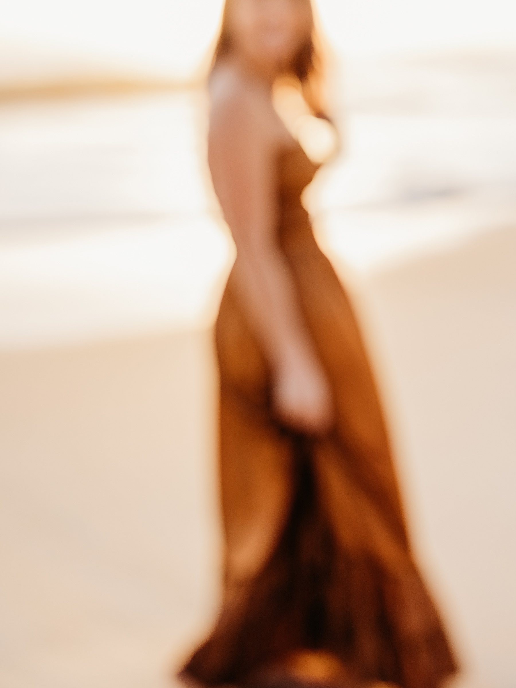 a blurry photo of a woman standing on the beach in a brown dress at sunrise
