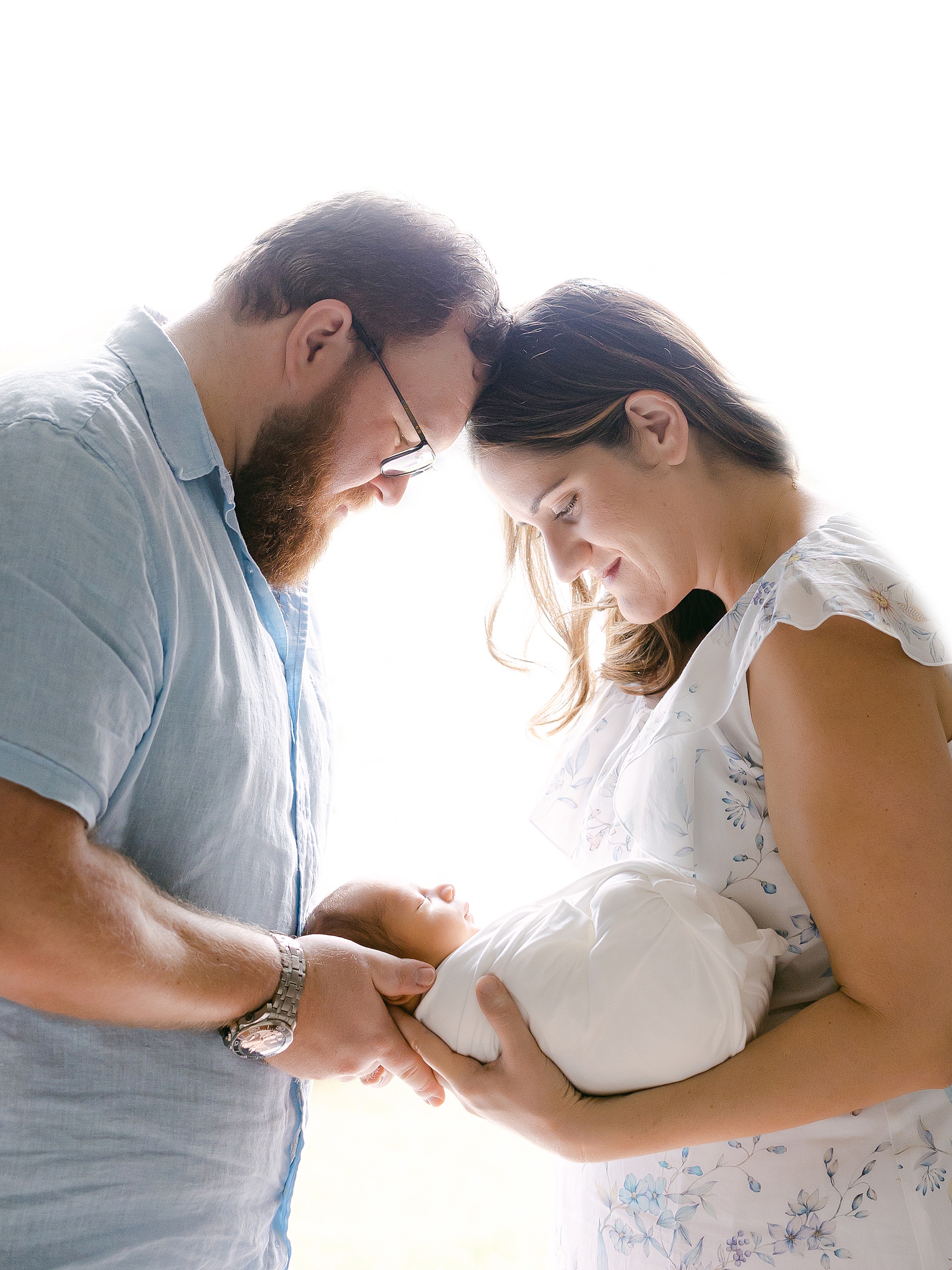 man and woman standing against window light holding newborn baby boy