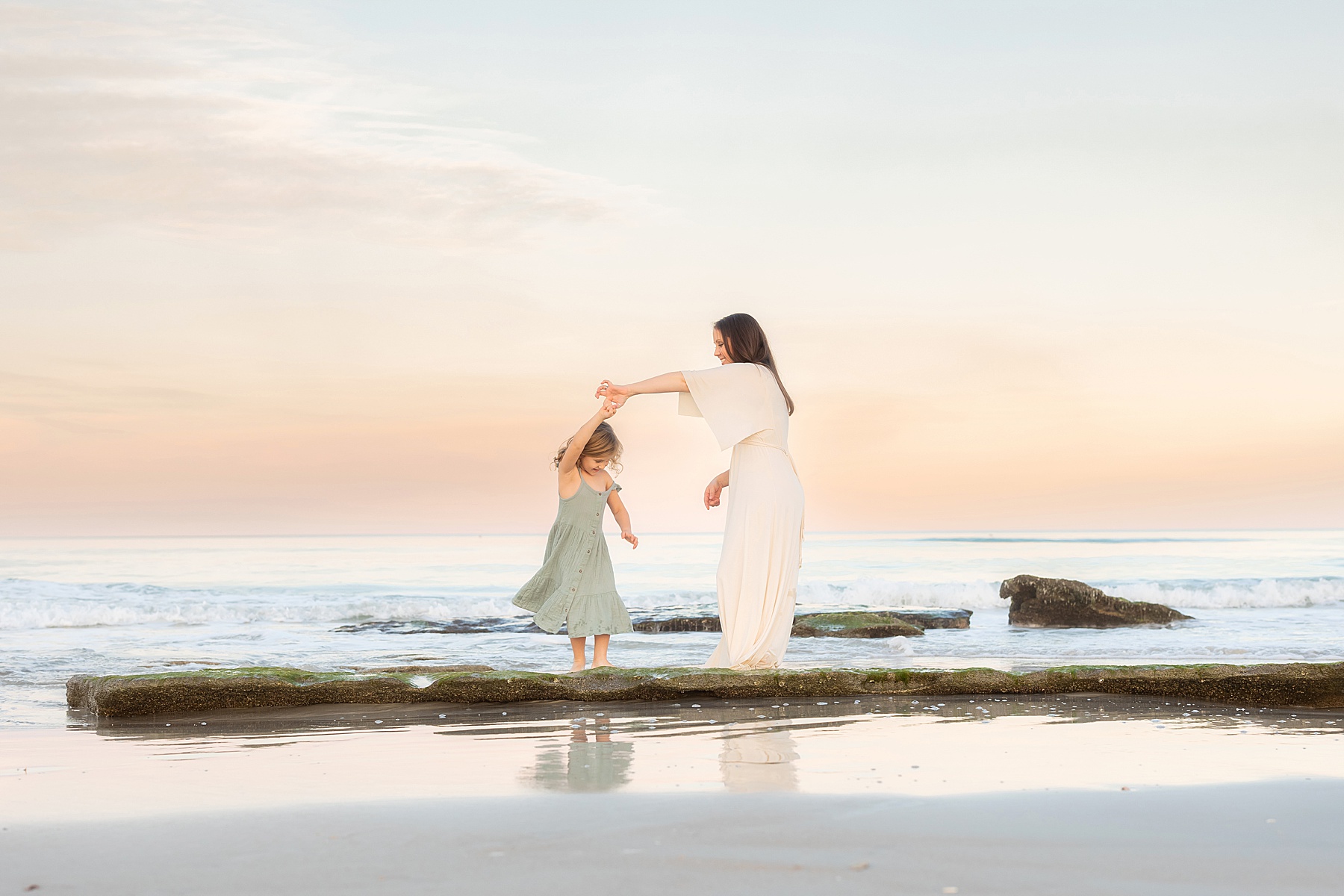 woman in cream dress standing on rock at the beach twirling little girl in a green dress