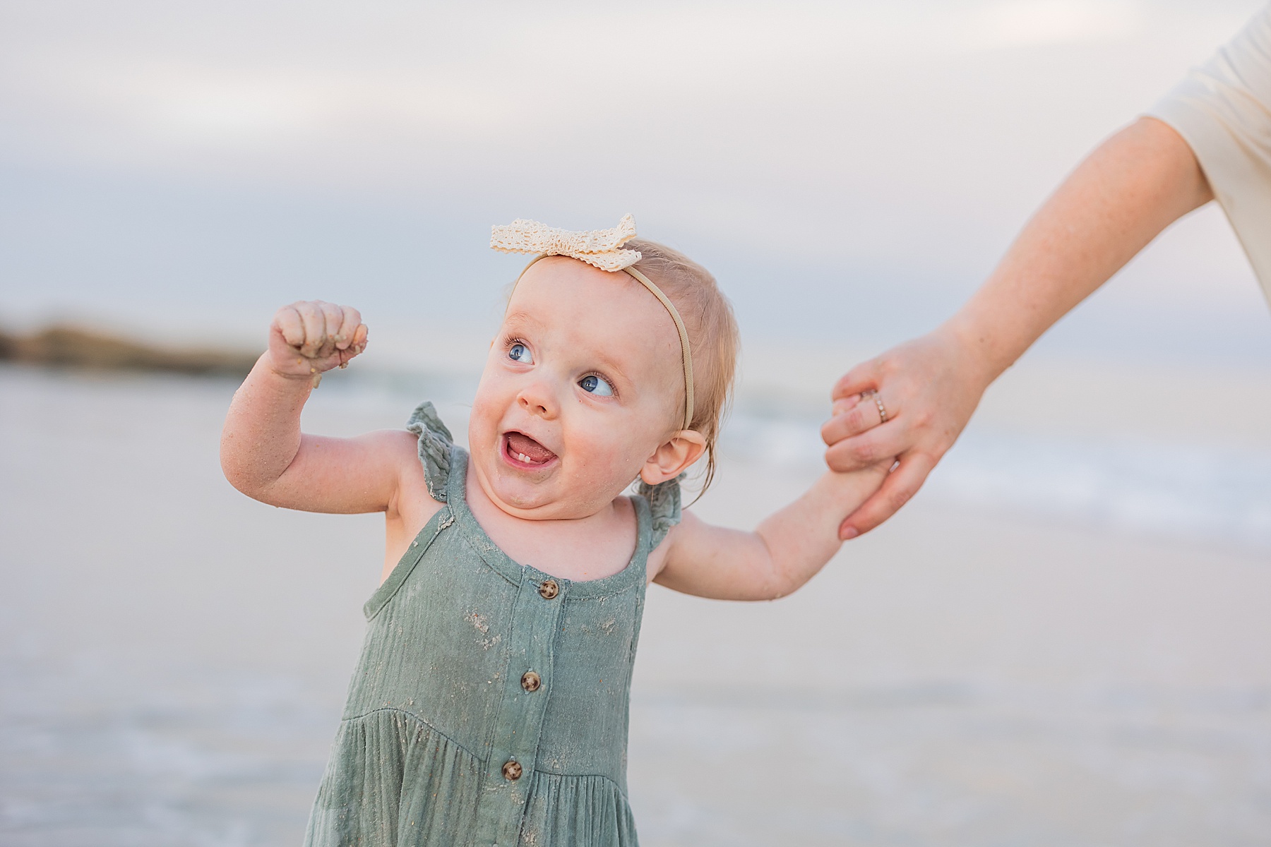 little girl in green dress making funny face holding her moms hand at the beach