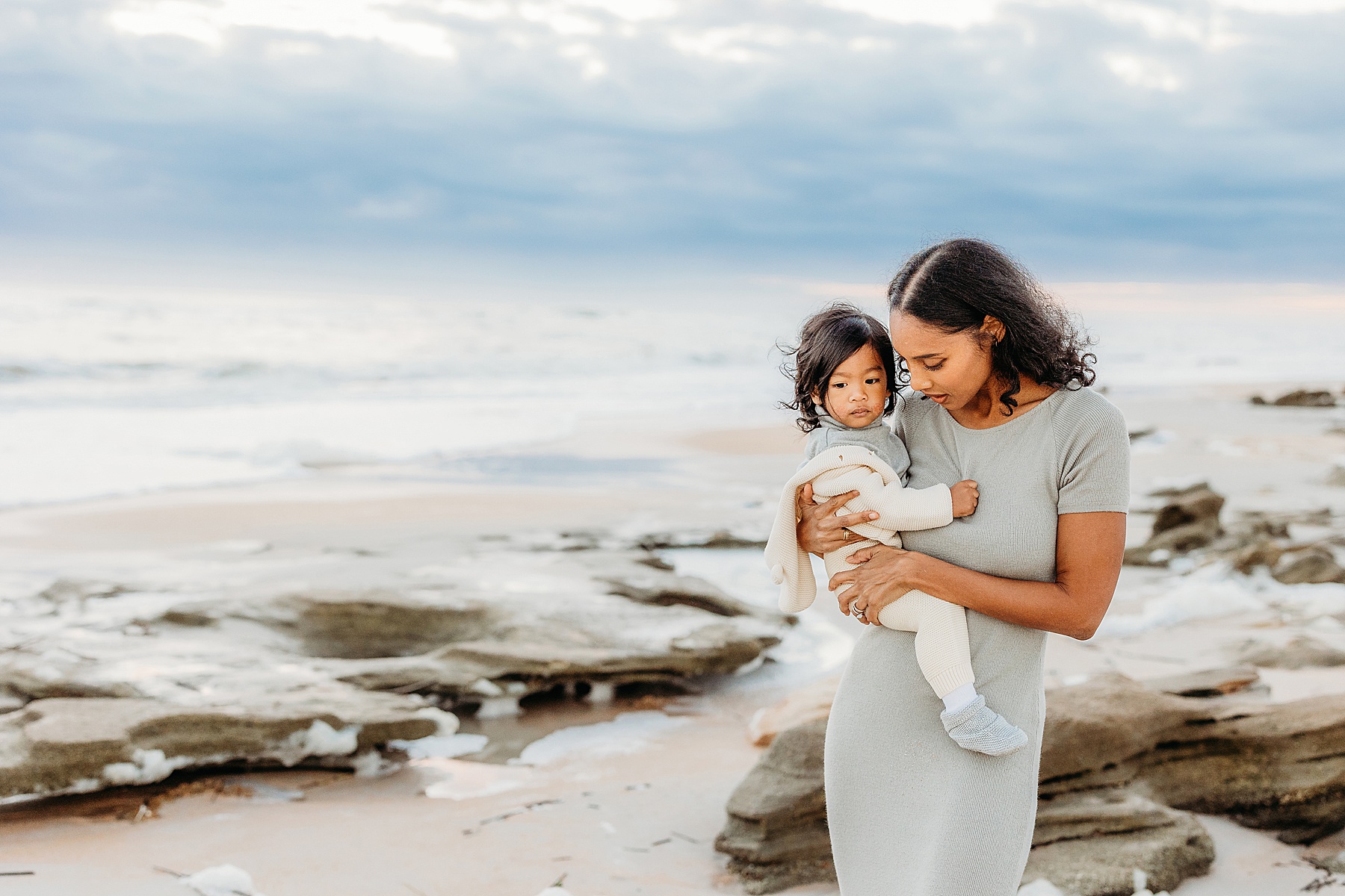 woman standing with baby boy on the beach at sunrise wearing a long gray dress