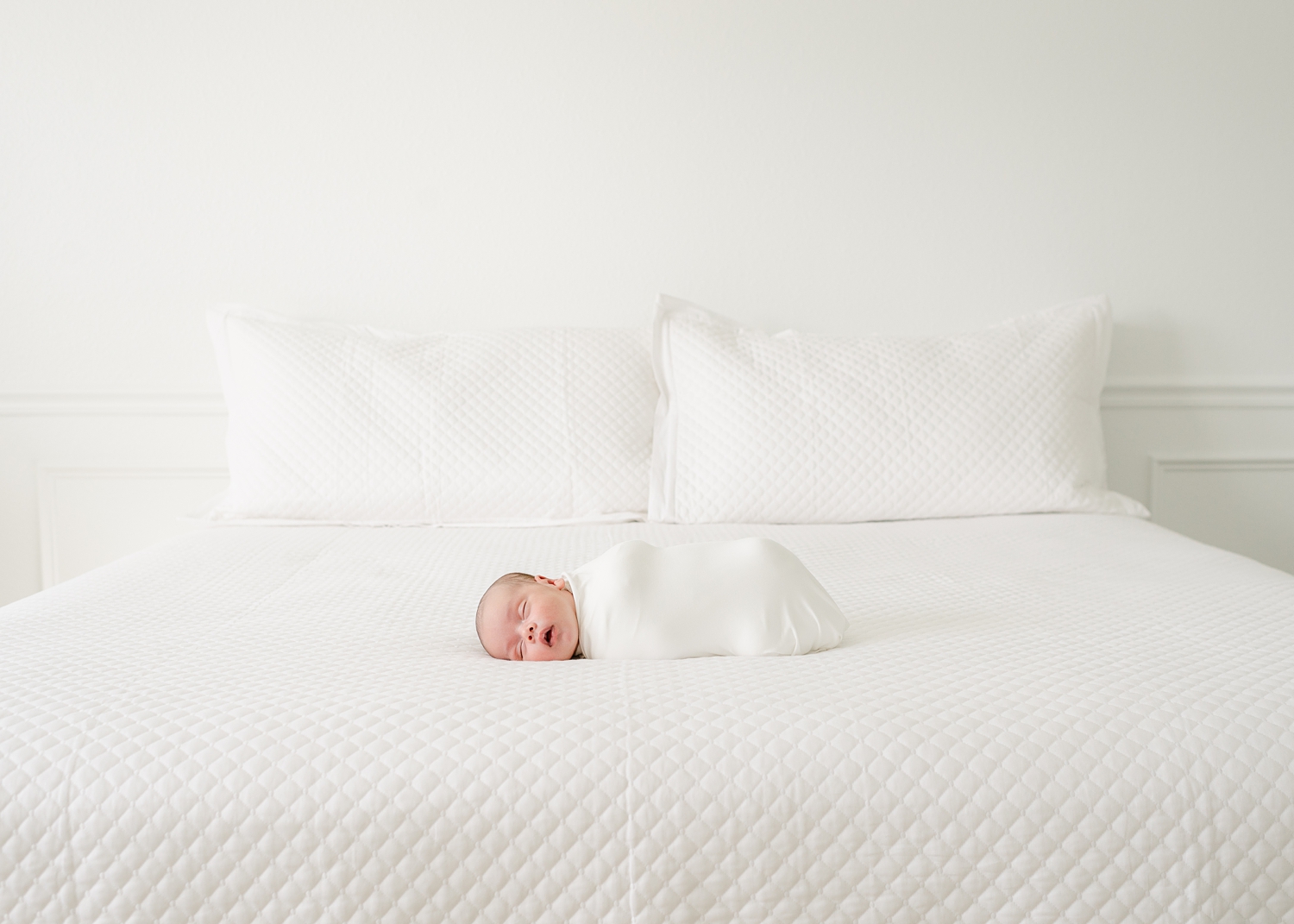 newborn baby boy wrapped in white blanket on all white bed sleeping