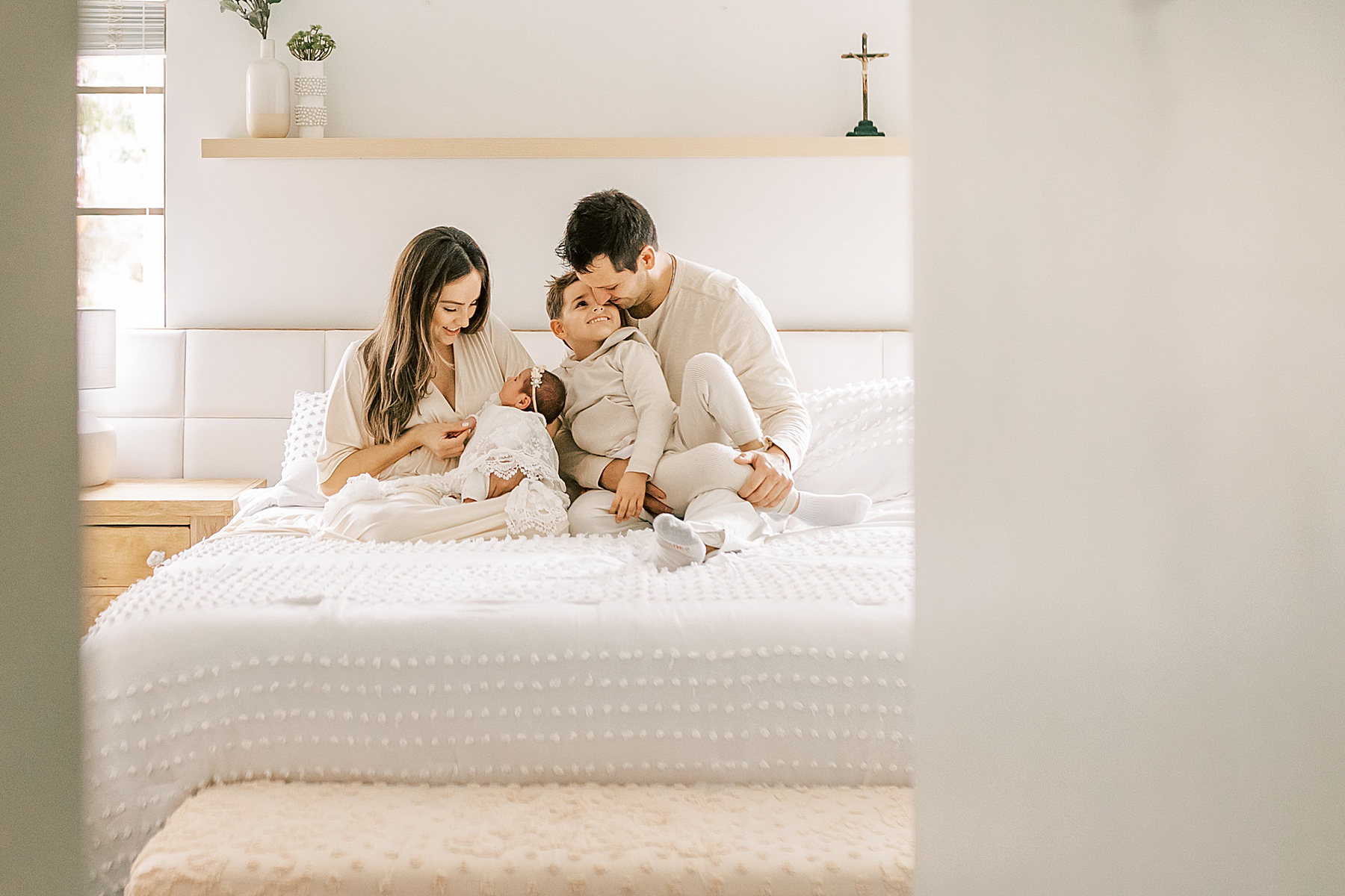 family dressed in neutral colors sitting on white bed looking at newborn baby girl