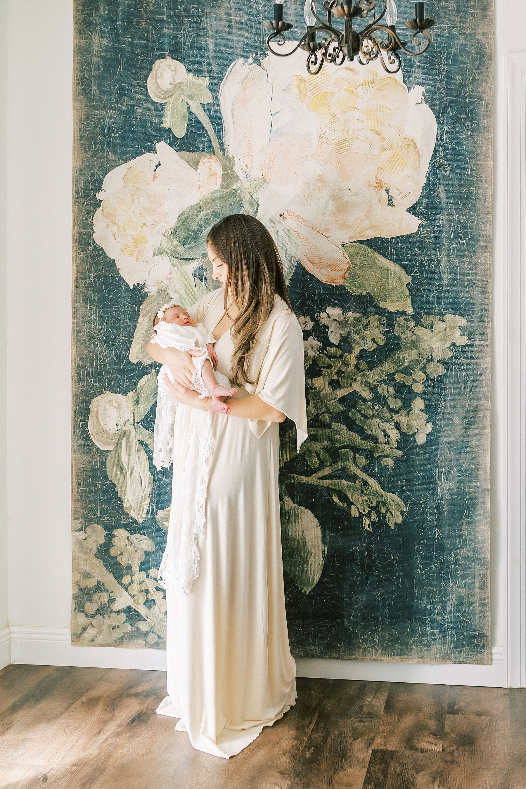 woman in cream dress with long dark hair in front of floral tapestry holding newborn baby girl wrapped in white lace wrap