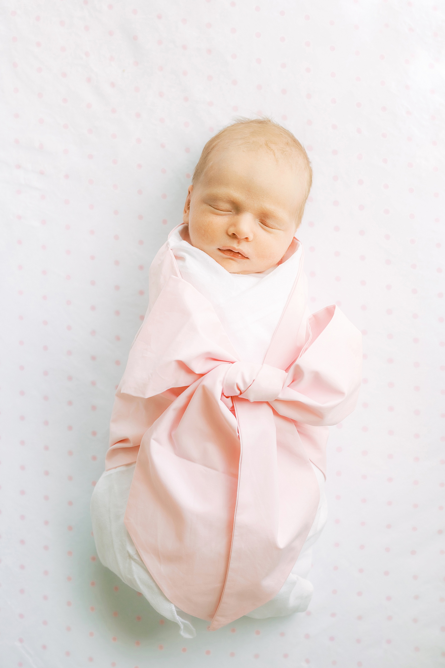 newborn baby girl wrapped in pink bow swaddle on white pink polka dot sheet