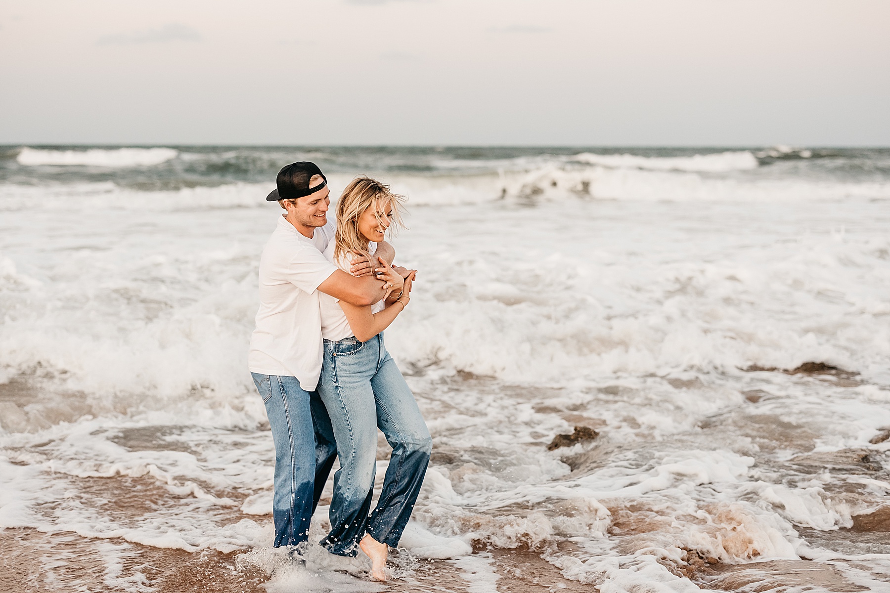 man and woman holding each other in the water on the beach wearing jeans and white shirts
